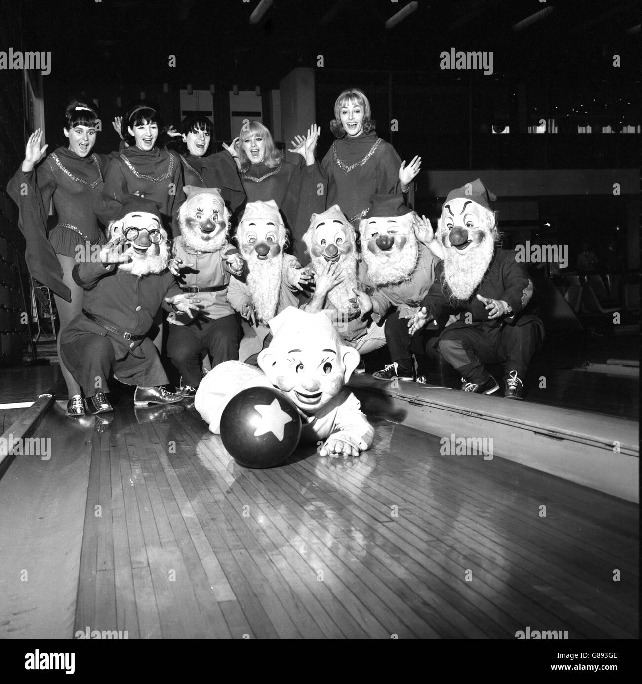 Ken Baker AS Dopey si lancia verso le spille al bowling a dieci birilli a Wembley, Londra, dove sta apparendo nella mostra di ghiaccio Snow White e i sette nani. Guardando sono i suoi sei compagni e alcune delle ragazze show. Foto Stock