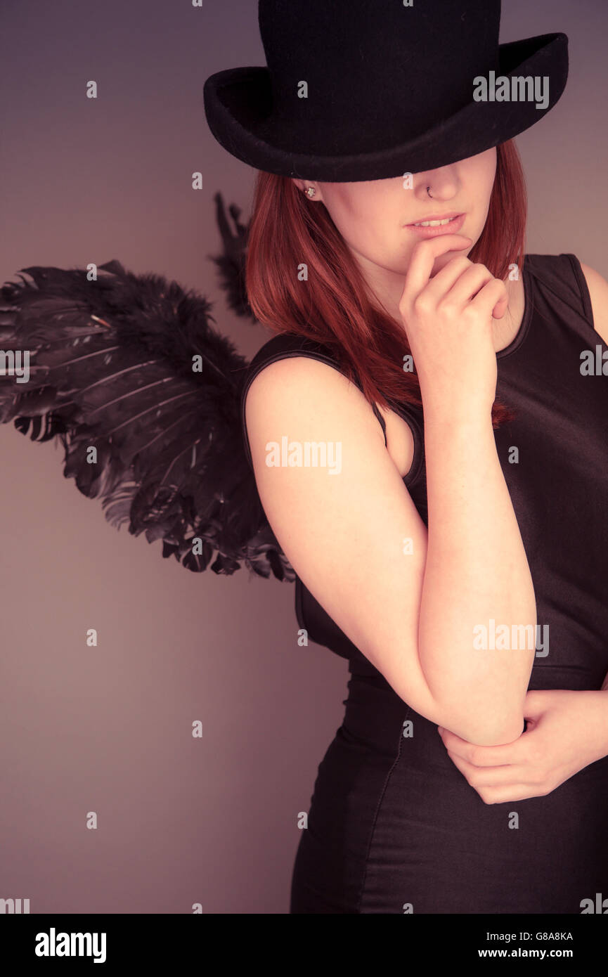 Un sottile giovani caucasici Ragazza donna che indossa un abito nero , un cappello che copre parzialmente il suo volto e una coppia di Dark Angel Wings cercando il pensiero contemplativo scrutando pensieroso, REGNO UNITO Foto Stock