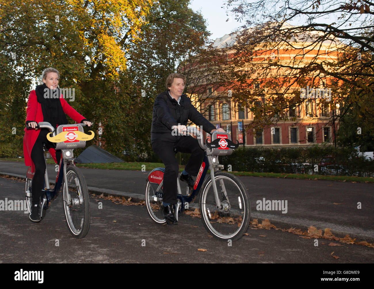 Katie Pugh (a sinistra) e Brandon Stockwell (a destra) sostenitori di Movember, pedala attraverso un parco mentre i baffi sono posti su Santander Cycles, il programma di noleggio biciclette di Londra, per sensibilizzare la Fondazione Movember, Kensington, Londra. Foto Stock