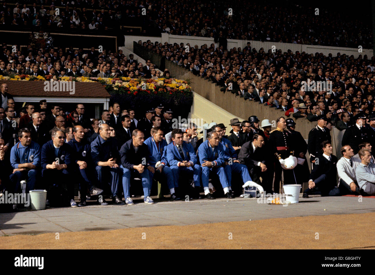 Le due panchine osservano la partita: (l-r) Dr H Schoberth della Germania occidentale, ?, assistente allenatore Dettmar Cramer, allenatore Helmut Schon e assistente allenatore Udo Lattek, e l'allenatore inglese Les Cocker, ?, direttore Alf Ramsey, fisico Harold Shepherdson e membro della squadra Jimmy Greaves. Sono anche presenti i membri della squadra inglese George Eastham (r), Ron Springett (secondo r), Peter Bonetti (terzo r), Ian Callaghan (appoggiato dietro Ramsey), Gerry Byrne (dietro Cocker) e Jimmy Armfield (davanti a Callaghan) Foto Stock
