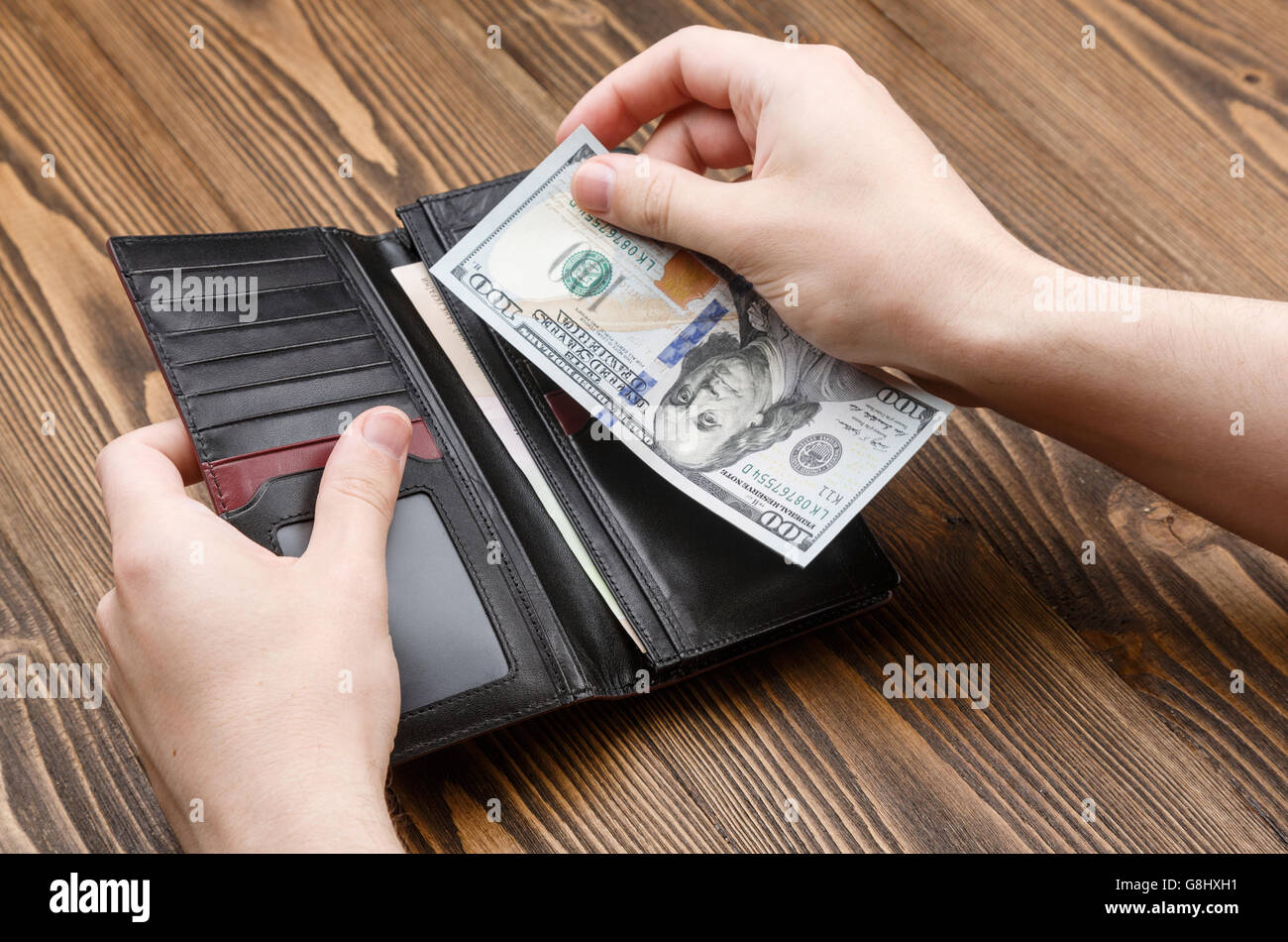 L uomo è messa 100 dollar banconota in un nero portafoglio in pelle su legno scuro dello sfondo. Immagine ravvicinata di mani e portafoglio. Foto Stock
