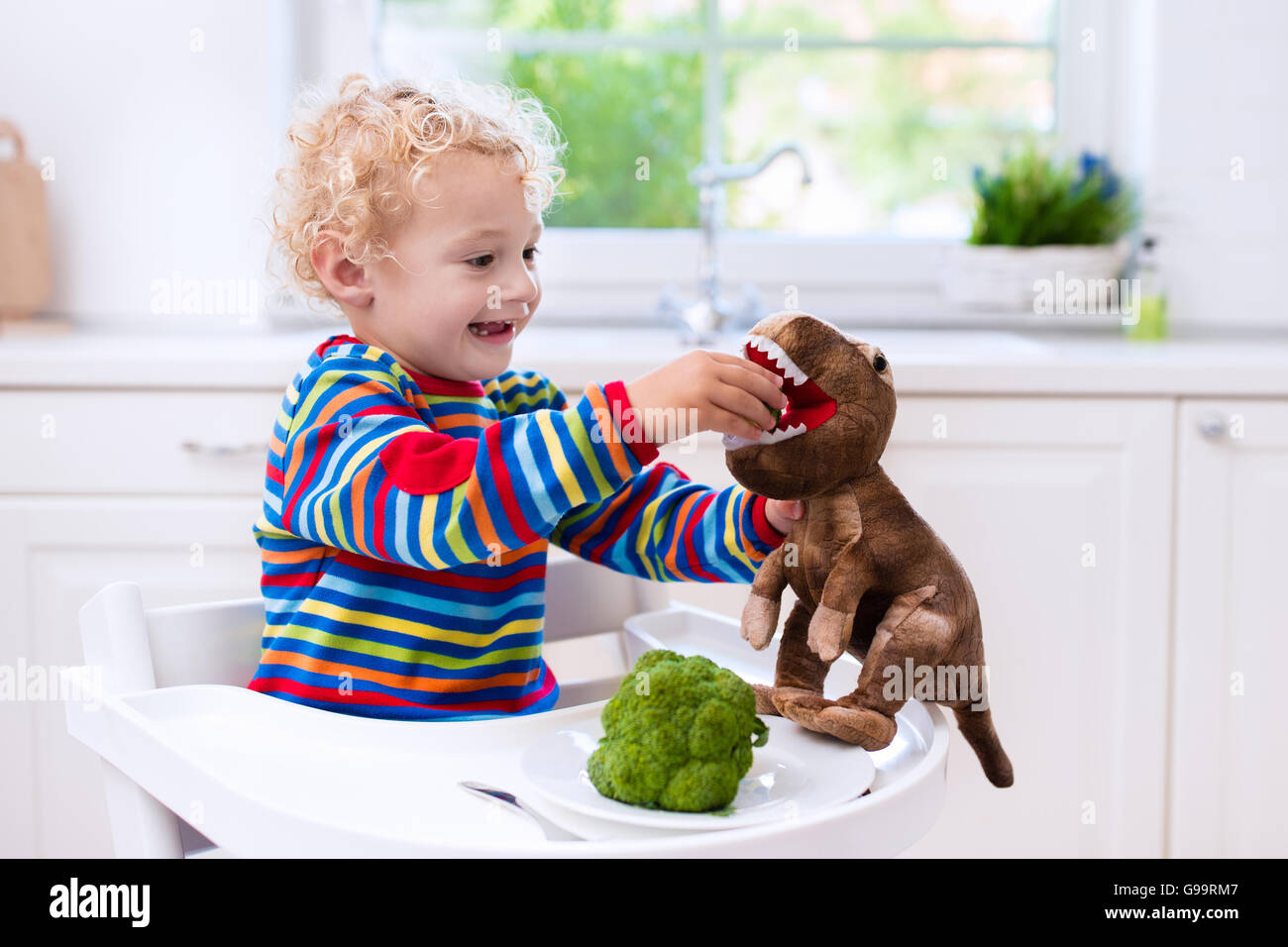 Happy Baby sitting in alta sedia di mangiare i broccoli in una cucina bianca. Una sana alimentazione per i bambini. Bio verdura come cibo solido Foto Stock