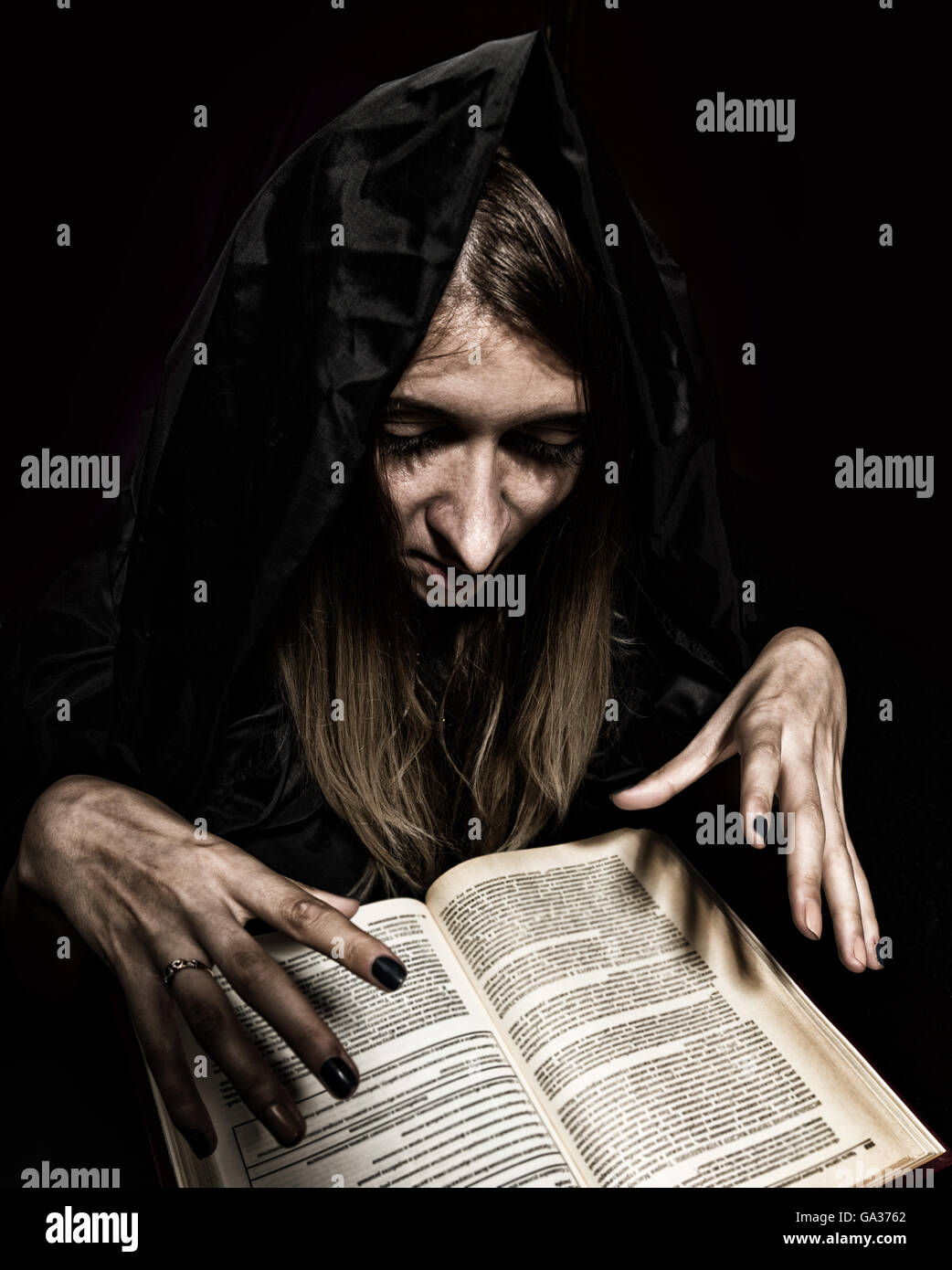 Bella strega getta incantesimi da spesso di libro antico a lume di candela su sfondo scuro Foto Stock