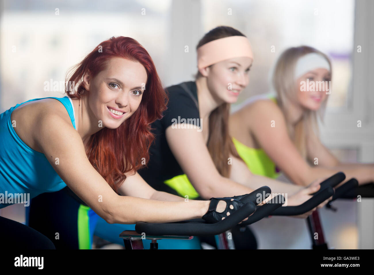 La pratica di fitness, gruppo di montare tre belle femmine allegro in bicicletta nel club sportivo, il riscaldamento durante lo sport lezione in classe Foto Stock