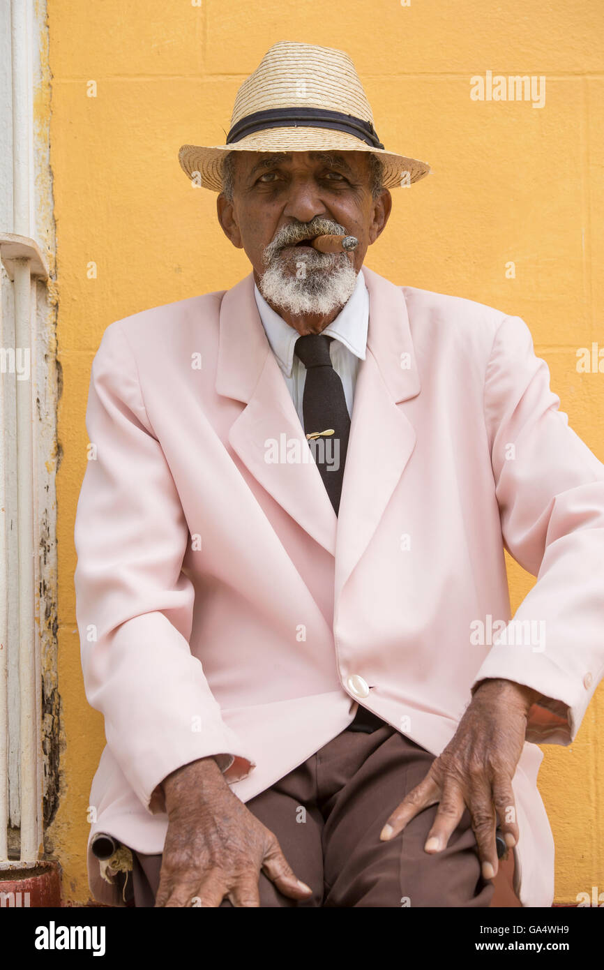 Ben vestito gentleman cubano in rosa blazer con cravatta seduti all'aperto di fumare un sigaro, Plaza Major, Trinidad, Cuba Foto Stock