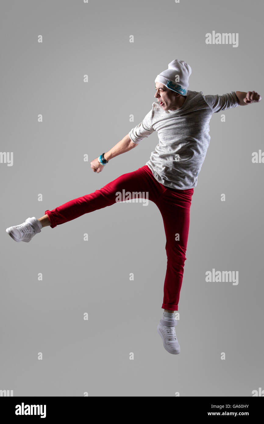 Giovani in stile moderno ballerino in casual pantaloni rossi che lavora fuori, ballando e saltando con divertenti grimace, cercando di kick qualcosa Foto Stock
