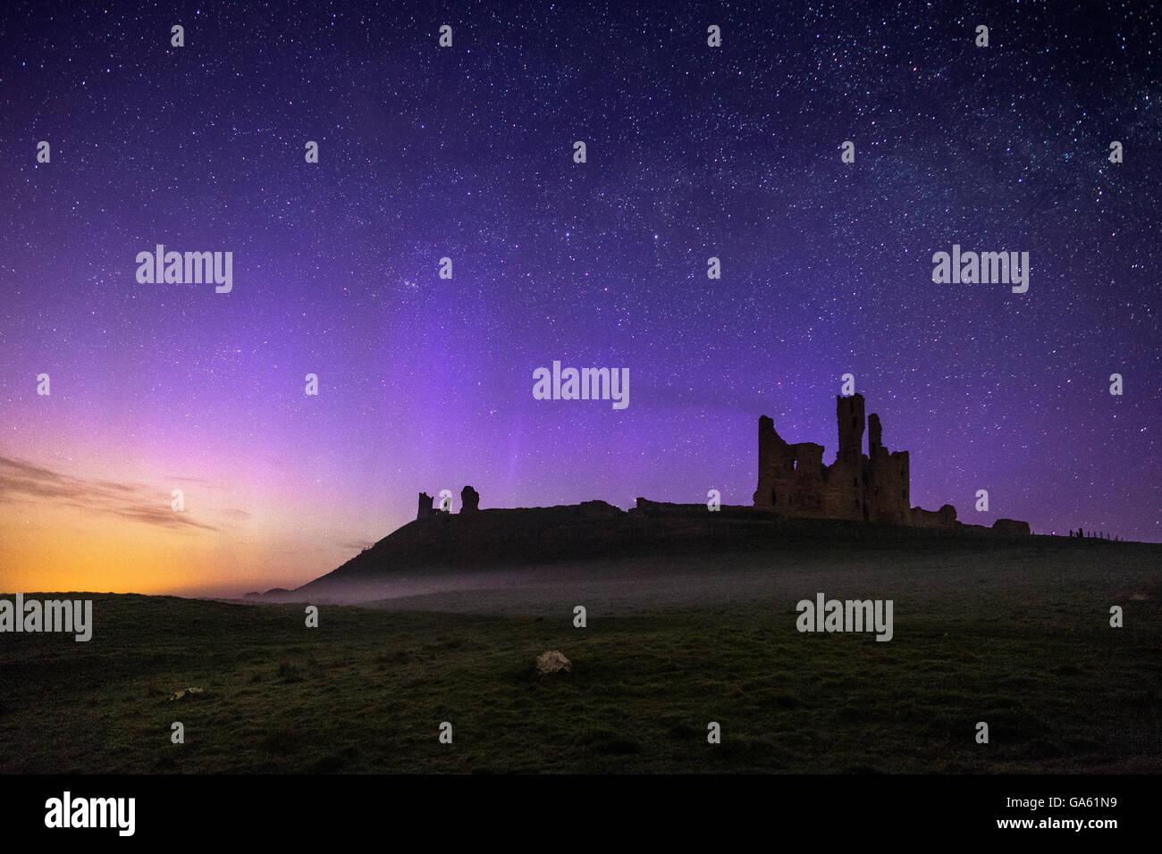 Dunstanburgh castello di notte con nebbia bassa e le luci del nord dancing across the sky. Foto Stock