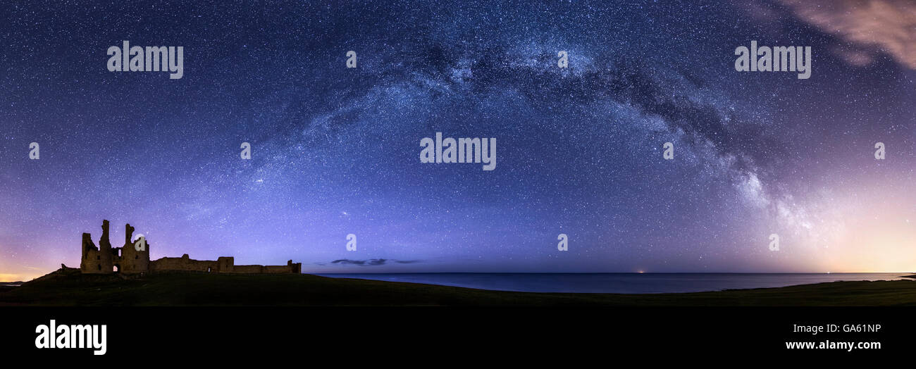 La Via Lattea emergente da un tripudio di inquinamento luminoso e la formazione di archi all'orizzonte verso il castello di Dunstanburgh in Northumberland Foto Stock