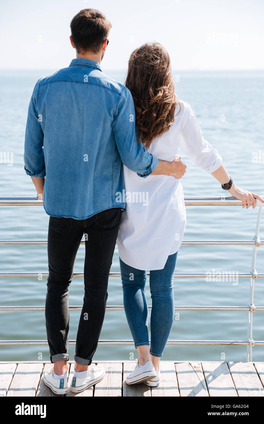 Lunghezza completa di una giovane coppia in piedi indietro abbracciando e guardando avanti al mare Foto Stock