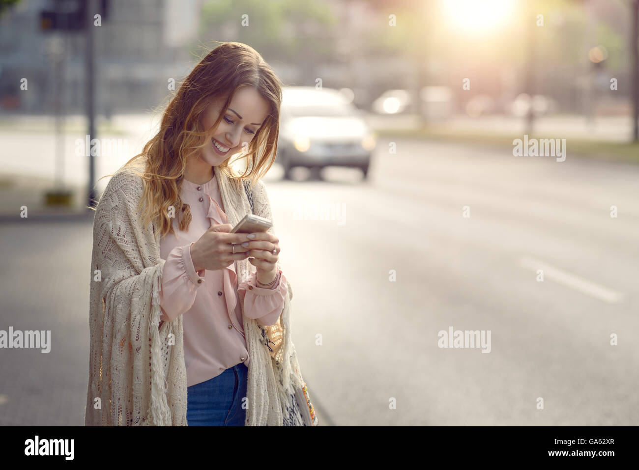 Attraente giovane donna controllo un messaggio sul suo telefono cellulare con un sorriso come essa si erge a lato di una strada urbana in Foto Stock