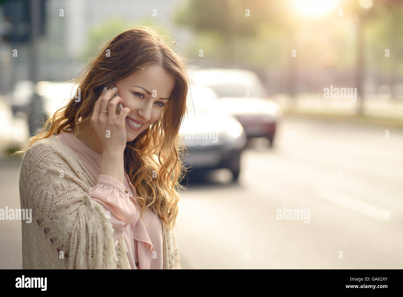Attraente giovane donna parlando sul suo telefono cellulare con un sorriso come essa si erge a lato di una strada urbana nei primi mesi del mattino Foto Stock