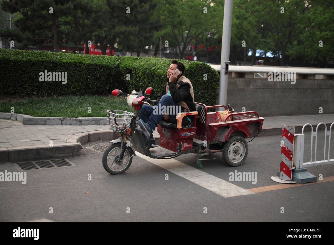 Un uomo sta parlando sul suo smartphone durante la guida di un triciclo motorizzato carrello. Donganmen Street, Pechino, Cina. 24.04.2016. Foto Stock