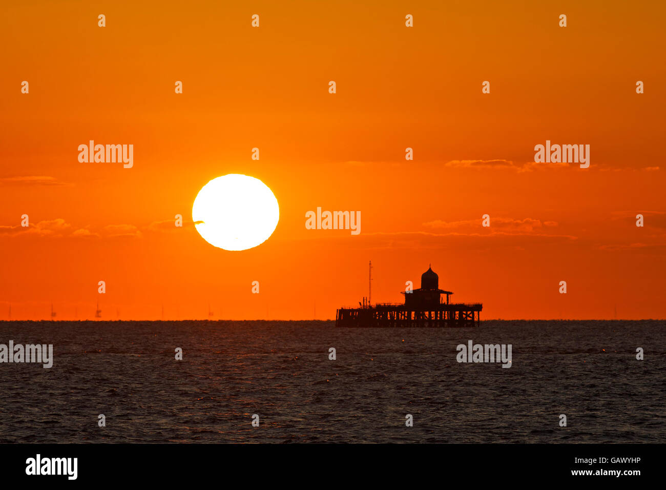 Herne Bay, Kent, Regno Unito. 6 Luglio 2016: Regno Unito Meteo. Sunrise dà il cielo di un rosso-arancione glow sopra i resti di Herne Bay pier, che era così gravemente danneggiato dalle tempeste nel 1978 che la sezione centrale è stata smantellata. Il London array, più grandi centrali eoliche offshore in tutto il mondo viene visto attraverso il calore haze. Temperature potrebbero raggiungere nella metà 20s durante il fine settimana come aria calda dal continente copre il sud del paese Credito: Alan Payton/Alamy Live News Foto Stock