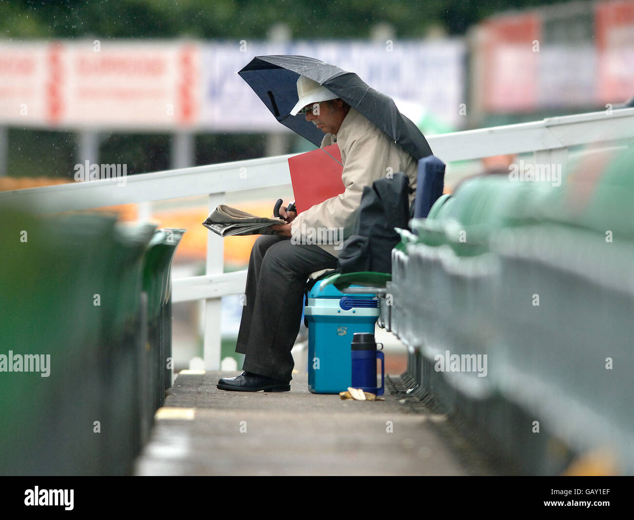 Mentre la pioggia comincia a cadere, uno spettatore si siede da solo nei supporti riempiendo in una crossword Foto Stock