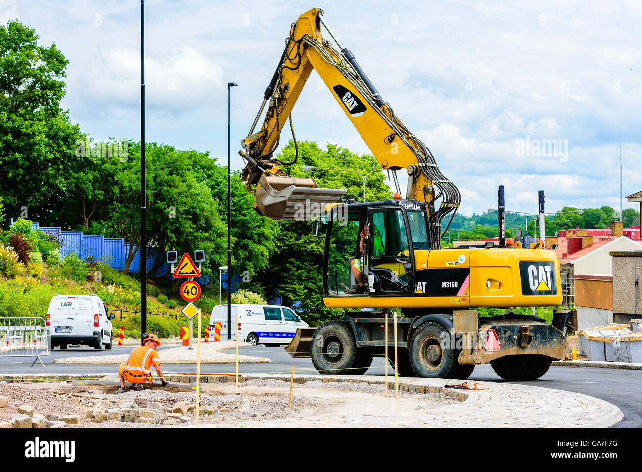 Motala, Svezia - 21 Giugno 2016: lavoratore di sesso maschile e di un escavatore CAT la costruzione di una rotatoria. Auto e segni di traffico in background. Rea Foto Stock