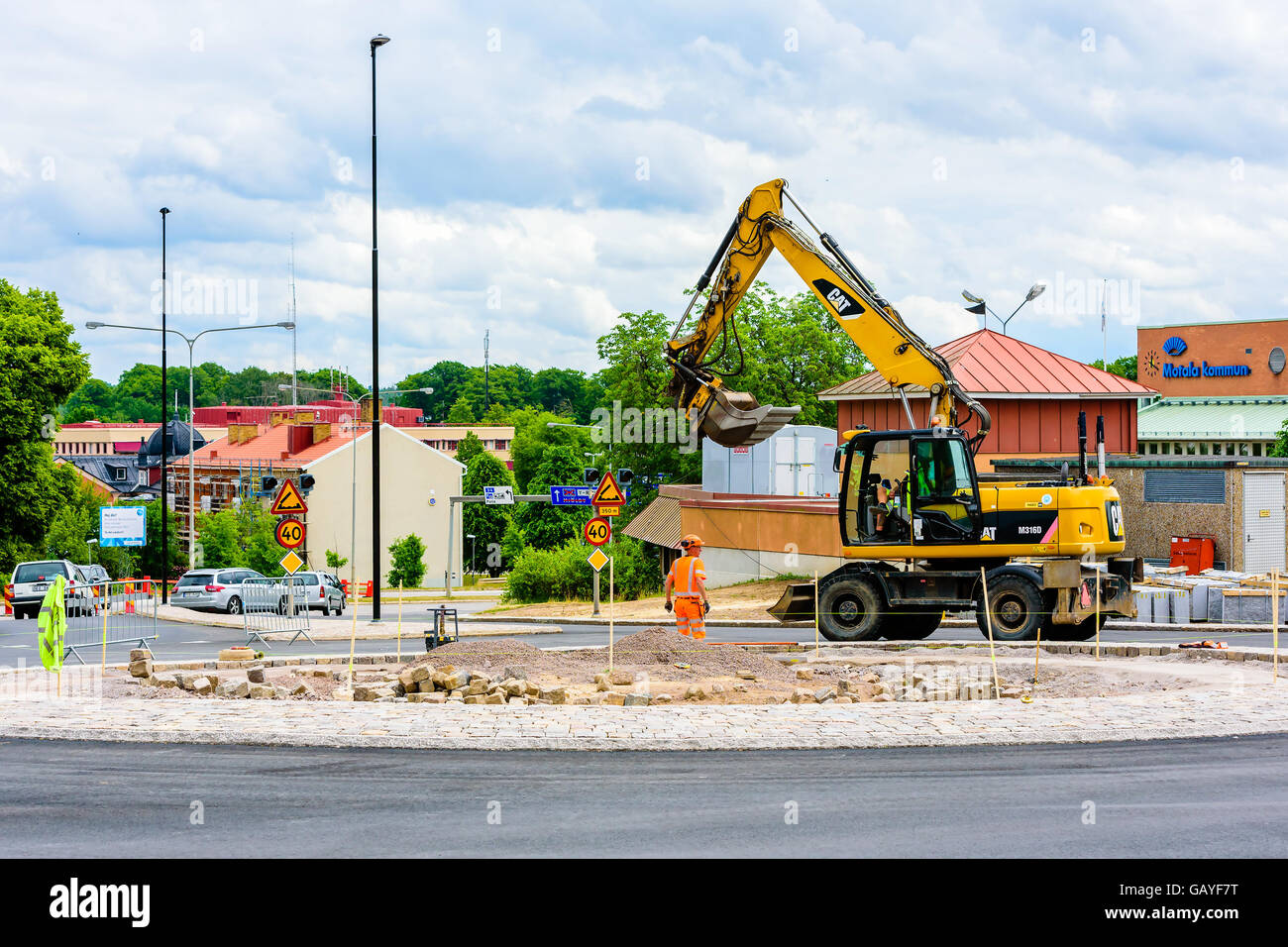 Motala, Svezia - 21 Giugno 2016: lavoratore di sesso maschile e di un escavatore CAT la costruzione di una rotatoria. Auto e segni di traffico in background. Rea Foto Stock