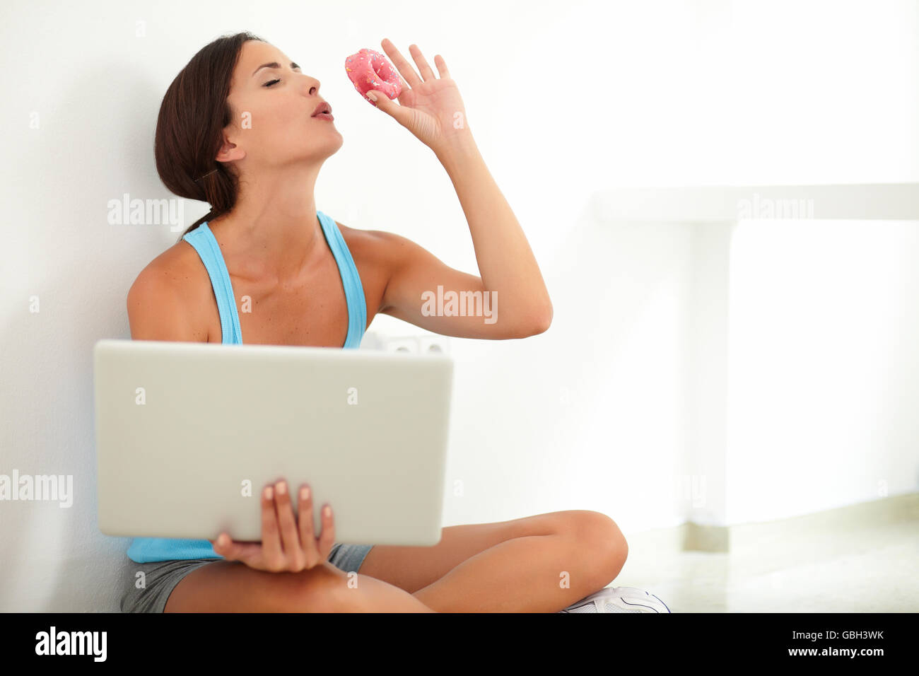 Bella femmina con gli occhi chiusi esplorando il web su computer e tenendo una ciambella mentre sogna in pausa la dieta Foto Stock