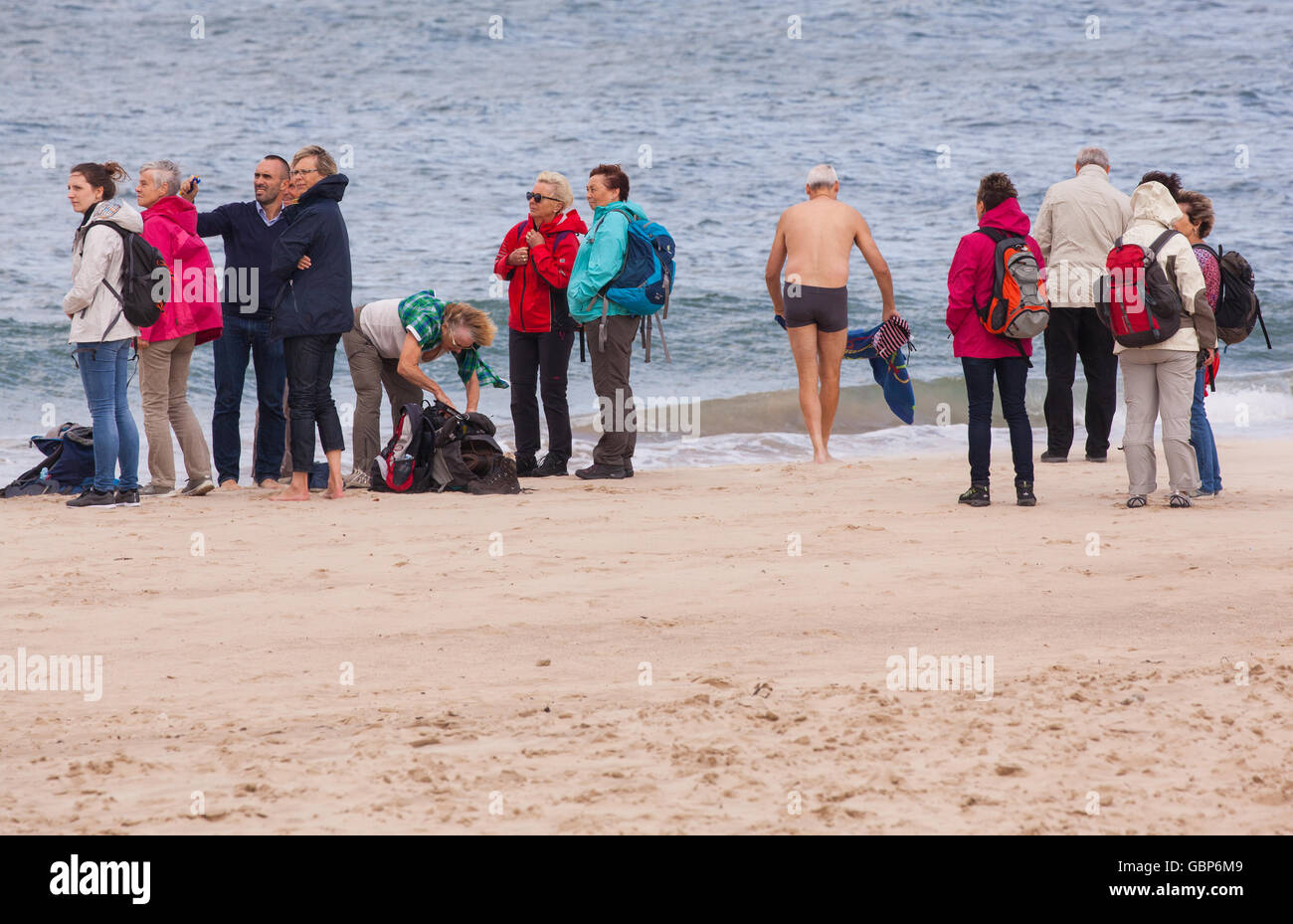 L uomo che sta per andare a nuotare in mare mentre il gruppo delle persone intorno a lui hanno tutti vestiti caldi e cappotti su Foto Stock