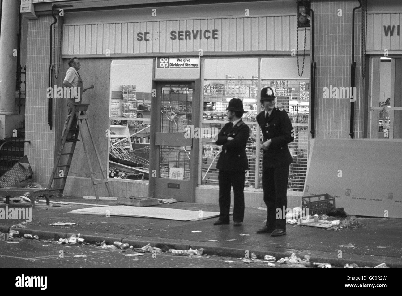 La guardia di guardia della polizia mentre un addetto si trova a salire su una finestra del Co-Op Supermarket e fuori licenza in Westbourne Park Road questa mattina dopo le scene violente della scorsa notte al carnevale di Notting Hill. Foto Stock