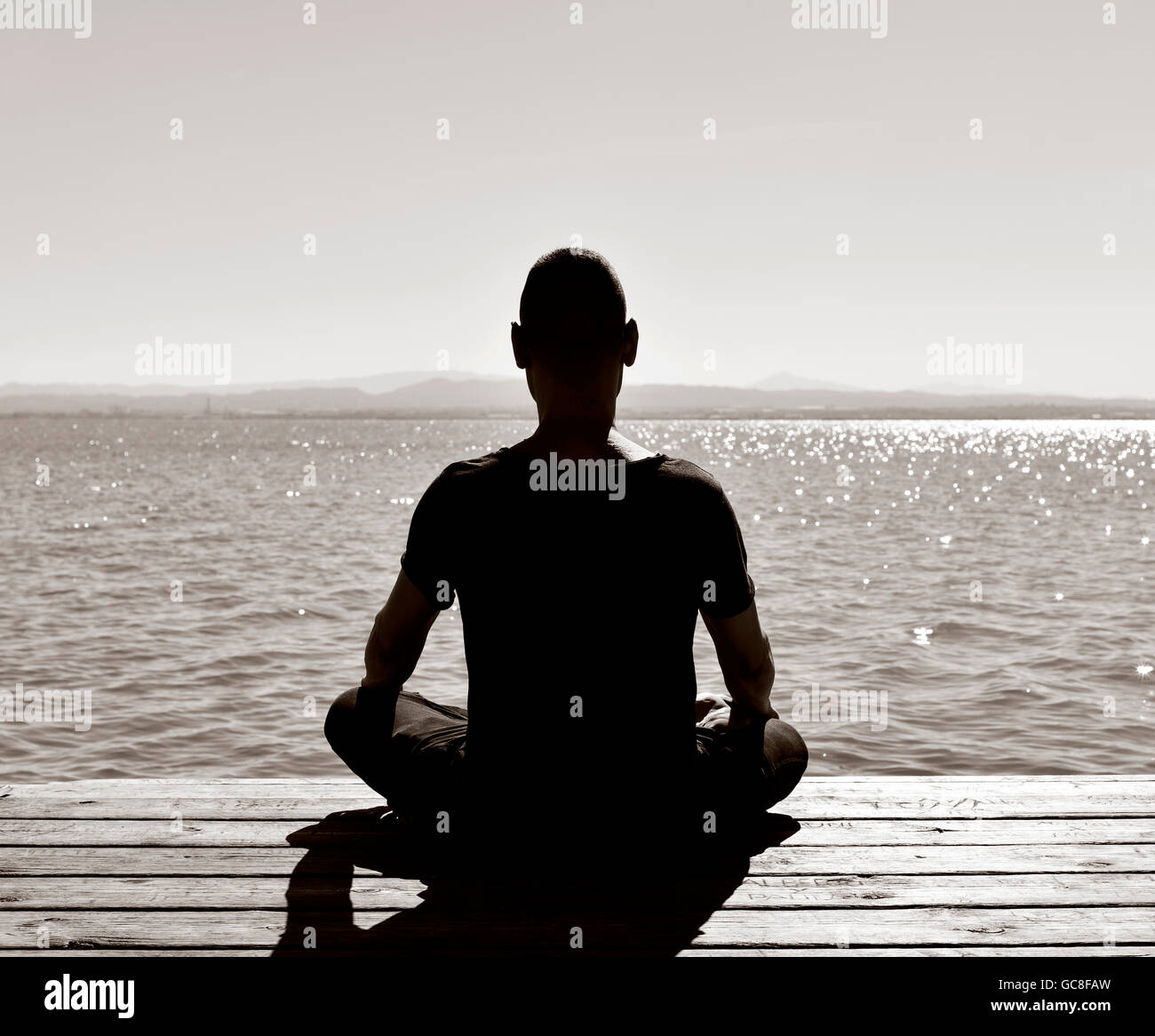 Giovane uomo visto da dietro meditando nella posizione del loto in un vecchio molo in legno sul mare o su un lago, in nero e di Pentecoste Foto Stock