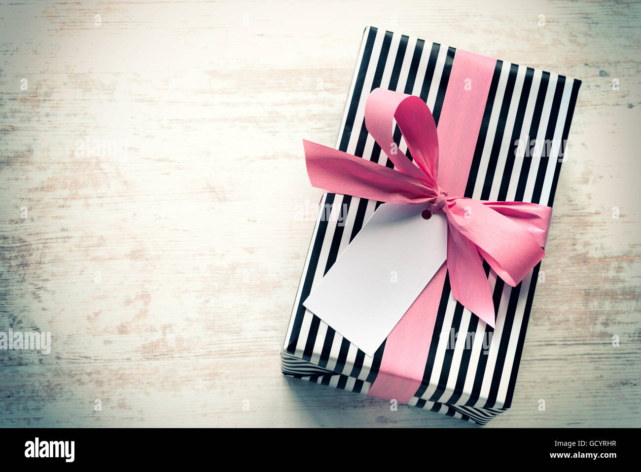 Confezione regalo avvolto in bianco e nero a strisce di carta con nastro rosa su di un legno bianco sfondo vecchio. Nota vuota legata al di sopra. Vintage Foto Stock