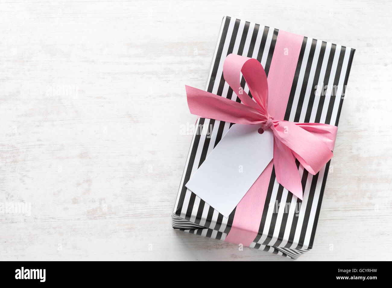 Confezione regalo avvolto in bianco e nero a strisce di carta con nastro rosa su di un legno bianco sfondo vecchio. Nota vuota legata al di sopra. Foto Stock