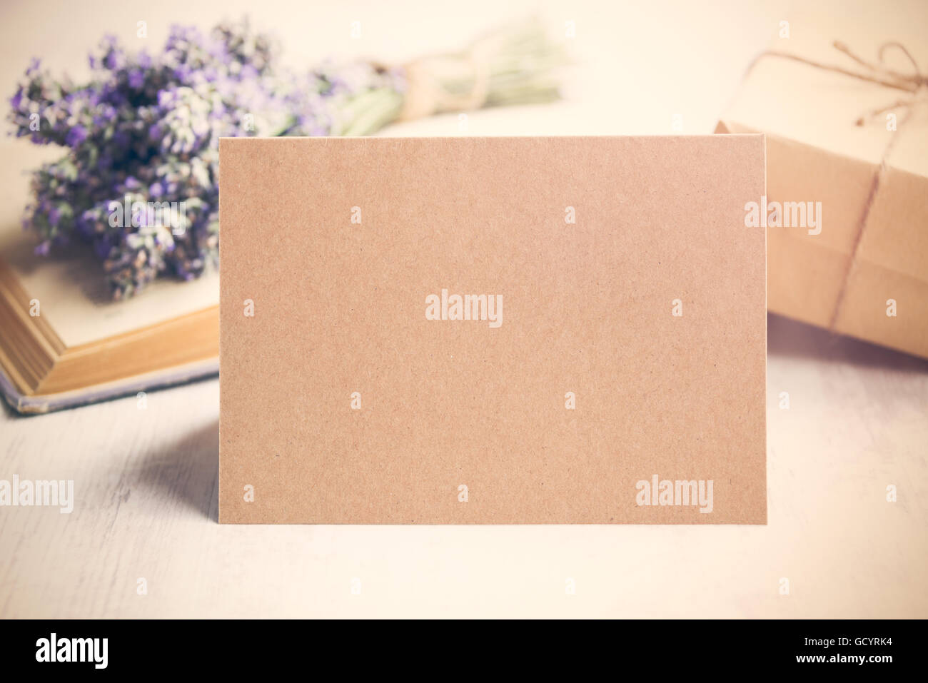 Saluto vuoto carta kraft nella parte anteriore di un mazzo di fiori di lavanda, avvolto un dono e un vecchio libro su un bianco sullo sfondo di legno. In stile vintage. Foto Stock