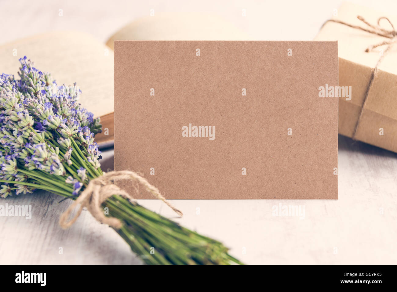 Saluto vuoto carta kraft nella parte anteriore di un mazzo di fiori di lavanda, avvolto un dono e un vecchio libro su un bianco sullo sfondo di legno. In stile vintage. Foto Stock