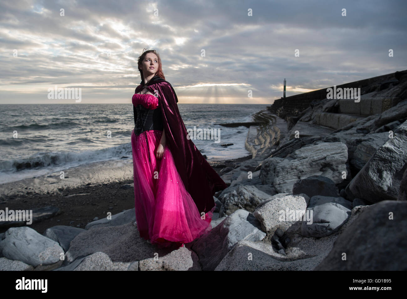 Outdoor " Moda goth' fantasy gotico make-over photo shoot con un dilettante non professionali di ragazza modello di donna che indossa un capo sulla posizione in piedi su una spiaggia rocciosa con un drammatico sky cloudscape dietro il suo Regno Unito Foto Stock