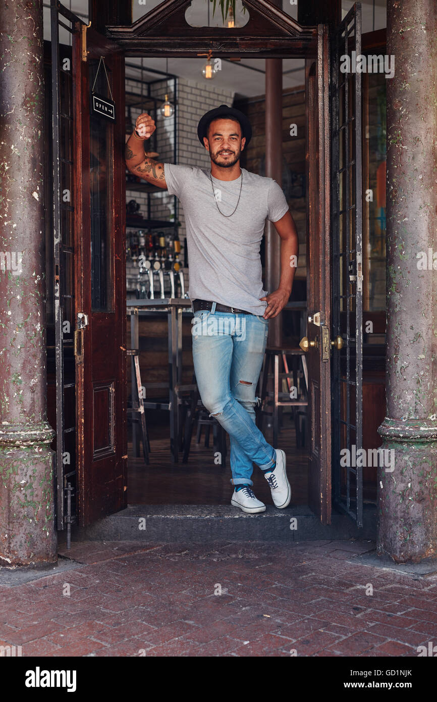 A piena lunghezza ritratto della moderna giovane maschio in piedi all'ingresso di un coffee shop. Elegante giovane a un cafe porta anteriore. Foto Stock