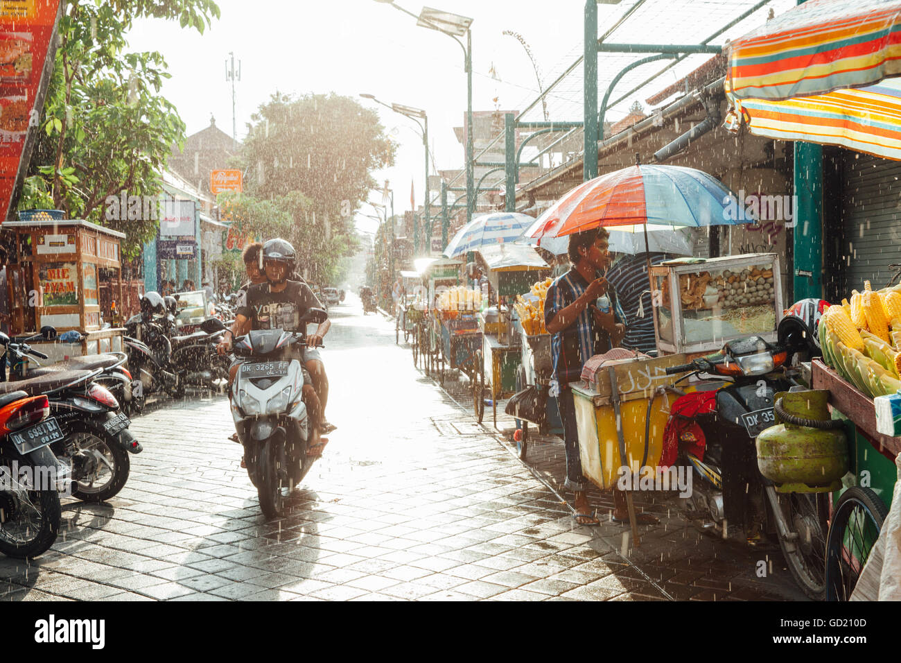 Kuta, Indonesia - 08 Marzo 2016: indonesiano fornitore alimentare nasconde dalla pioggia sotto ombrello della sua pressione di stallo sulla strada di Kuta. Foto Stock