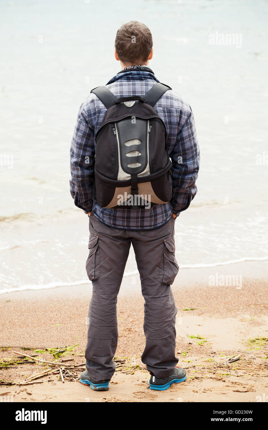 Traveler sul Mar Baltico. Giovane adulto uomo caucasico in caldo abbigliamento outdoor con zaino con protagonista al mare, vista posteriore Foto Stock
