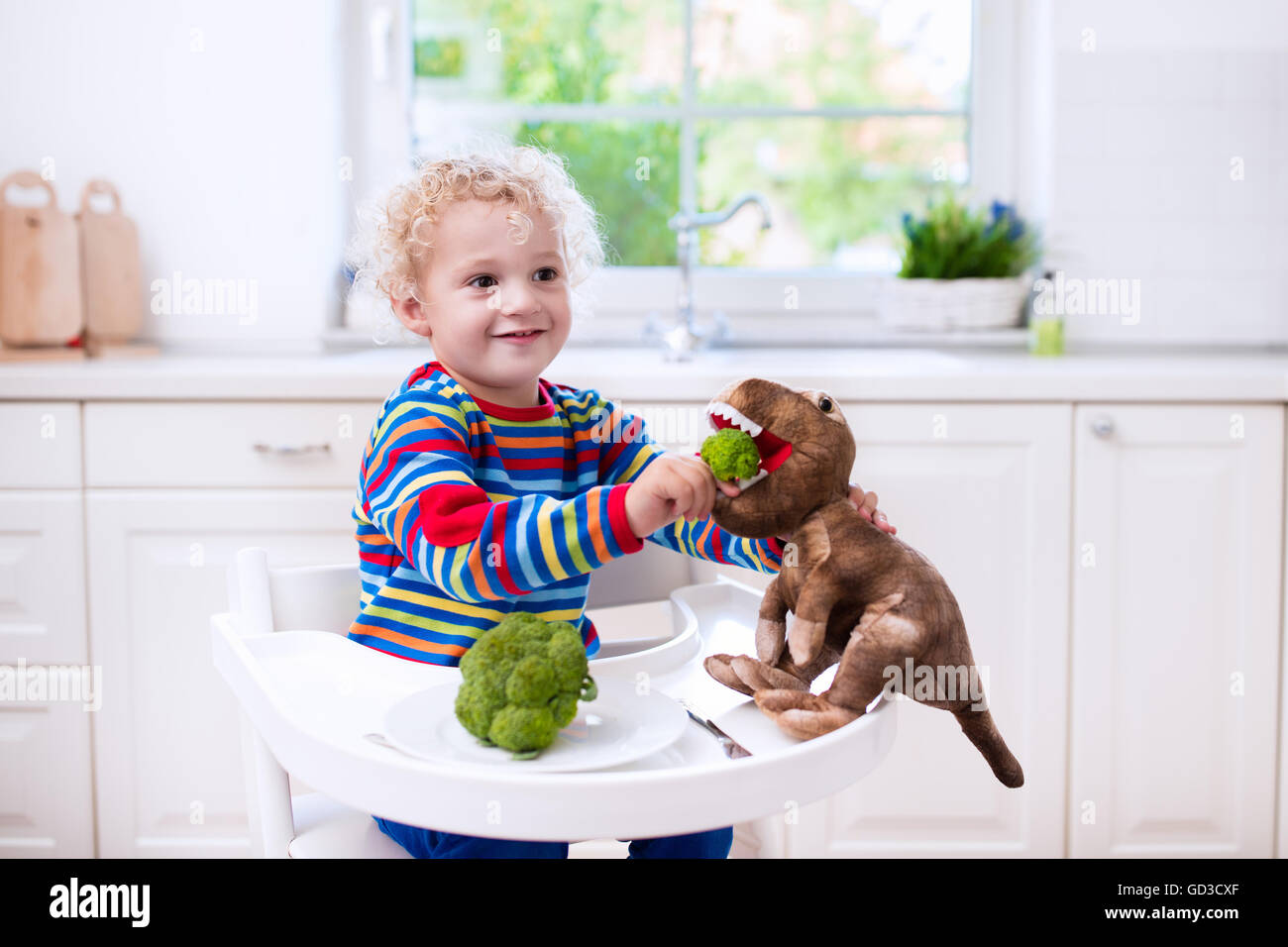 Happy little boy in sedia alta mangiare broccoli e alimentando il suo dinosauro giocattolo in una cucina bianca. Foto Stock