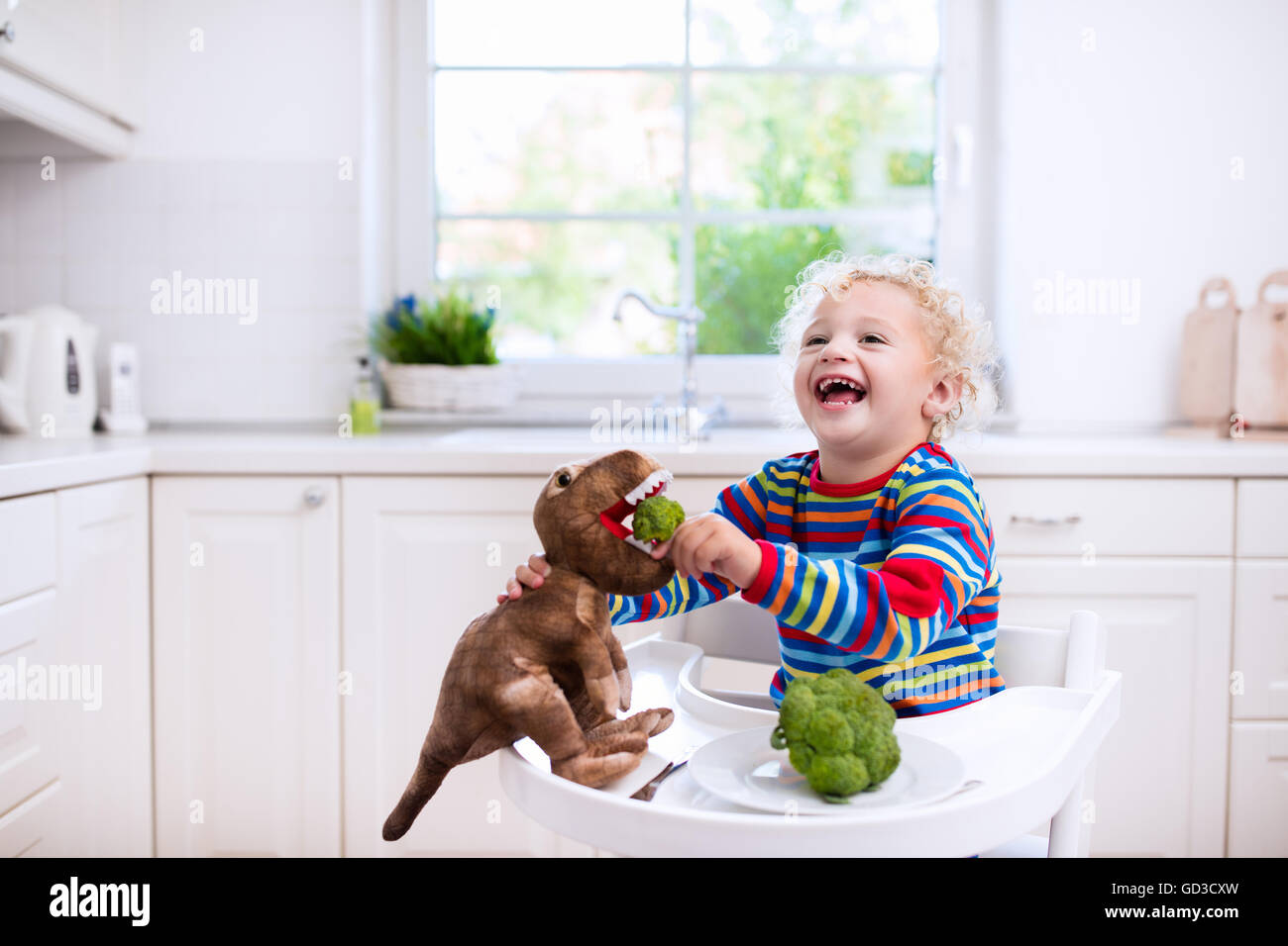 Happy little boy in sedia alta mangiare broccoli e alimentando il suo dinosauro giocattolo in una cucina bianca. Foto Stock