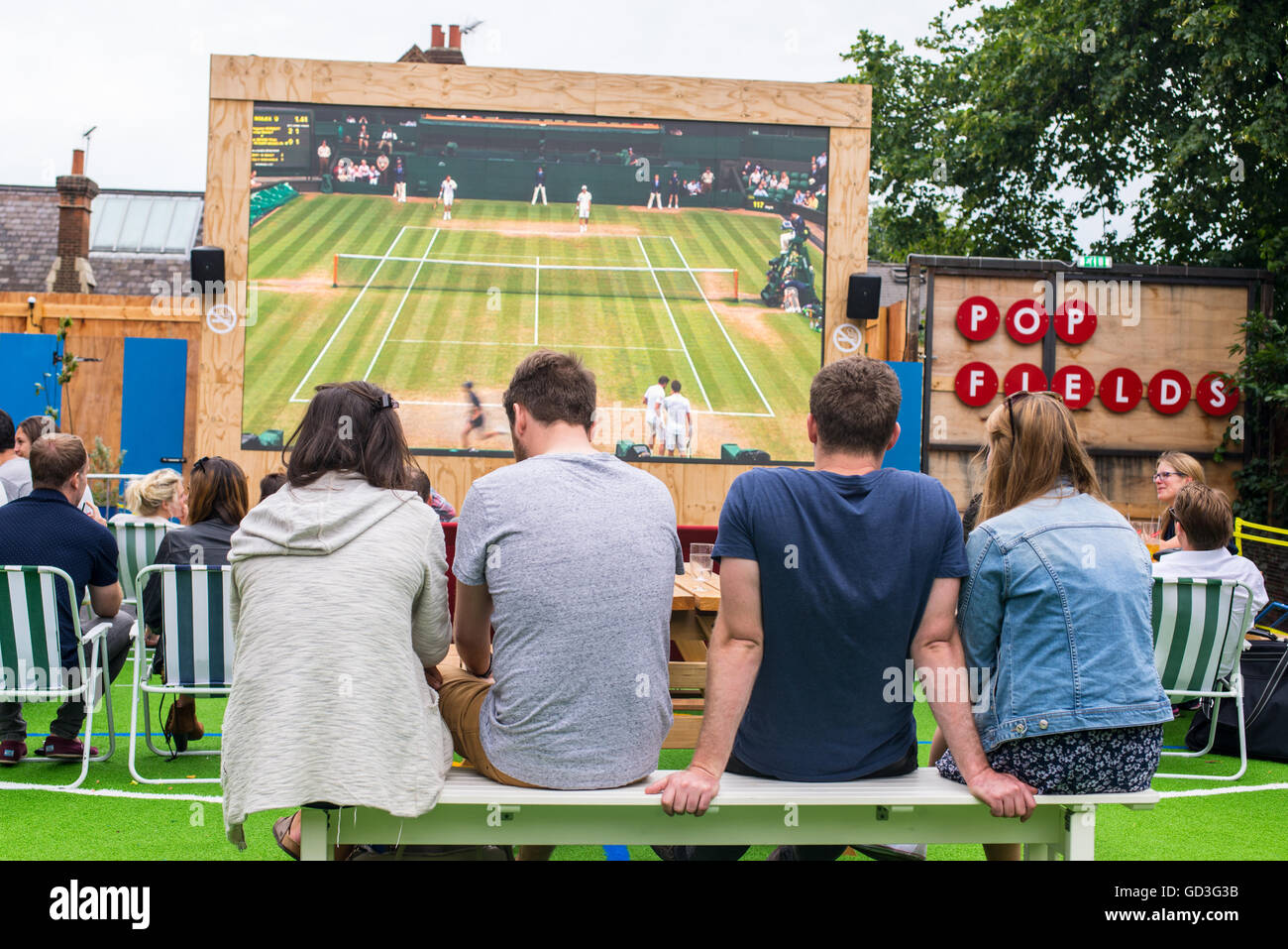 Persone prese da dietro la visione di un torneo di Wimbledon match di tennis nel grande schermo per esterni nei campi pop, parte del luogo alla moda Pop Brixton Foto Stock