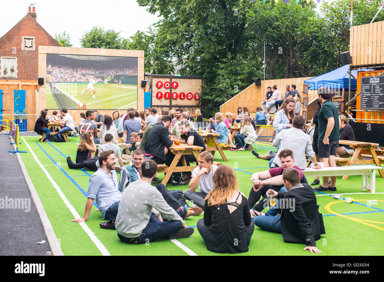 Persone che parlano e bere durante la visione di Wimbledon Tennis match in un grande schermo per esterni nei campi pop, Brixton Foto Stock