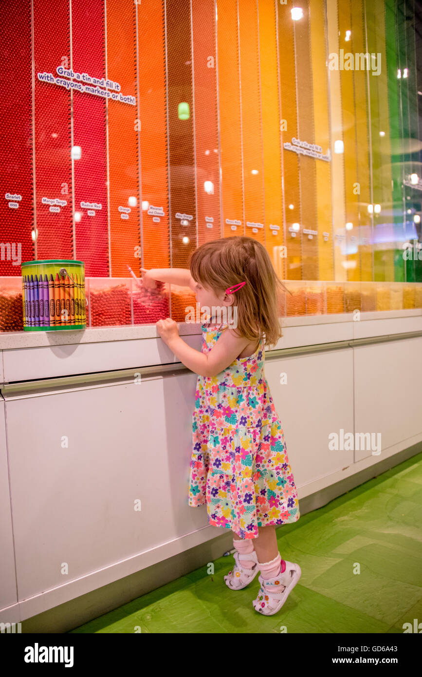 Bambina in piedi in punta di piedi in una colorata arte negozio di fornitura per la selezione di cera colorata matite colorate da un display con strisce di parete Foto Stock
