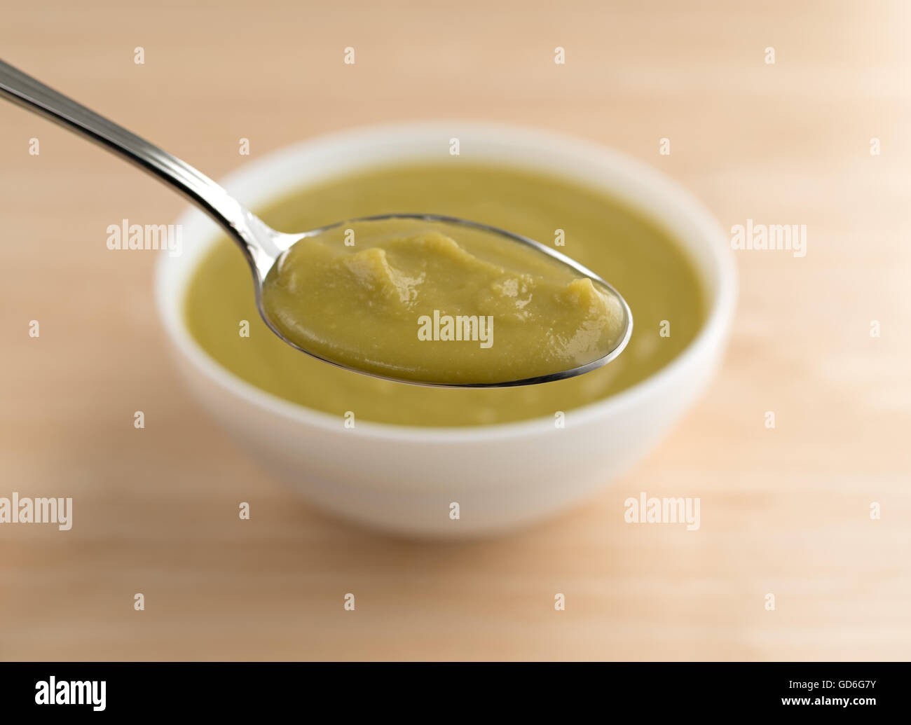 Un cucchiaio di verde spessa zuppa di piselli in primo piano con una vaschetta bianca in background su una tavola di legno alto. Foto Stock