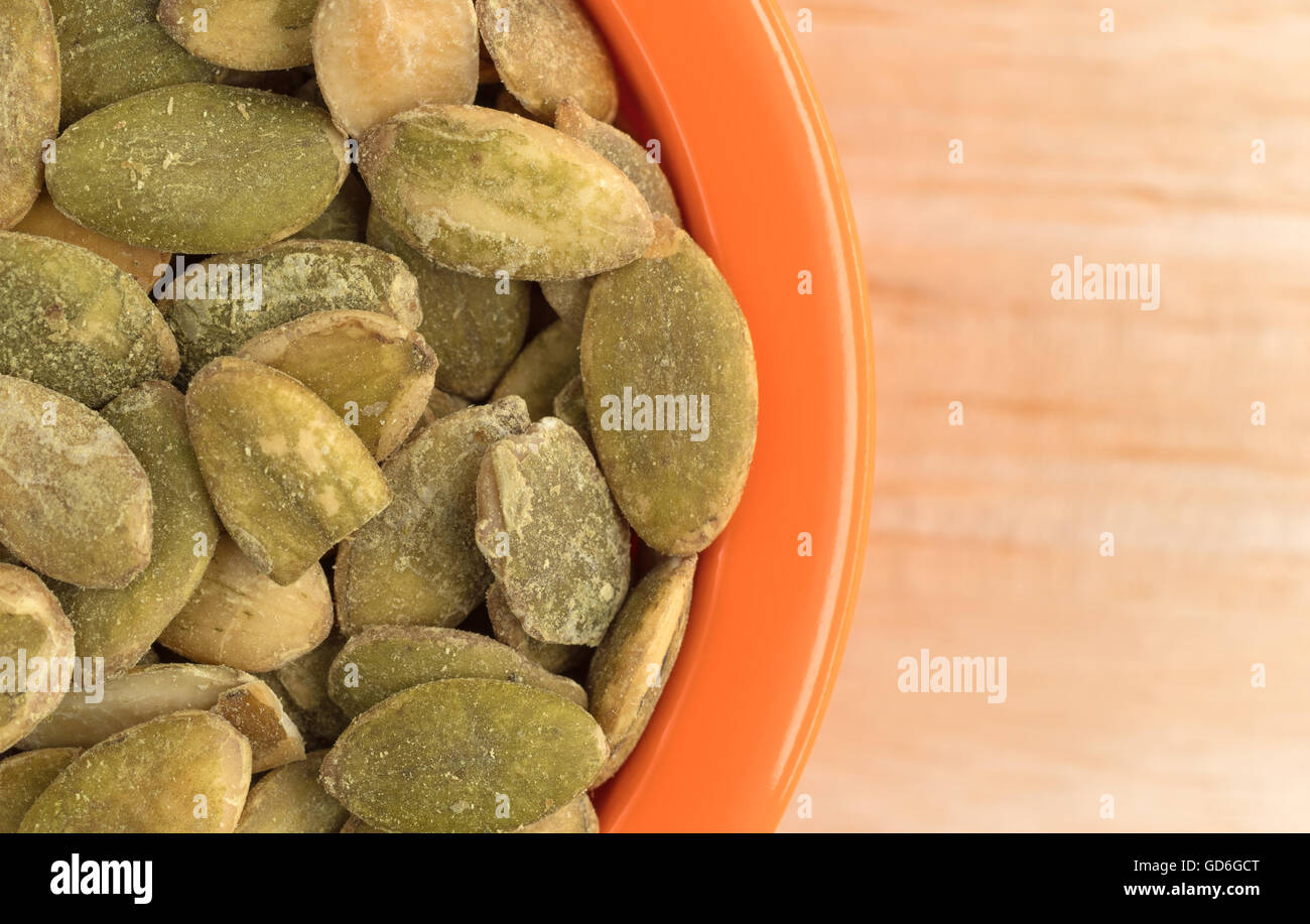 Top vista ravvicinata di una piccola ciotola arancione riempito con organici asciugare i semi di zucca su una tavola di legno alto. Foto Stock