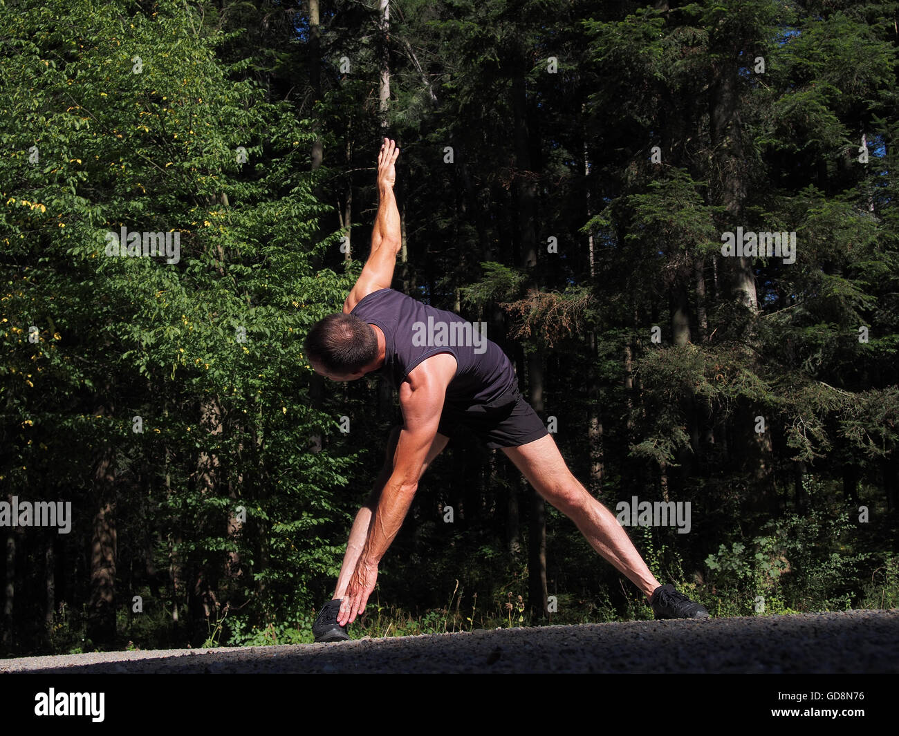 Maschio caucasico runner atleta stretching all'aperto nella foresta. Ampia zampe avanti permanente di piegare con torsione. Foto Stock