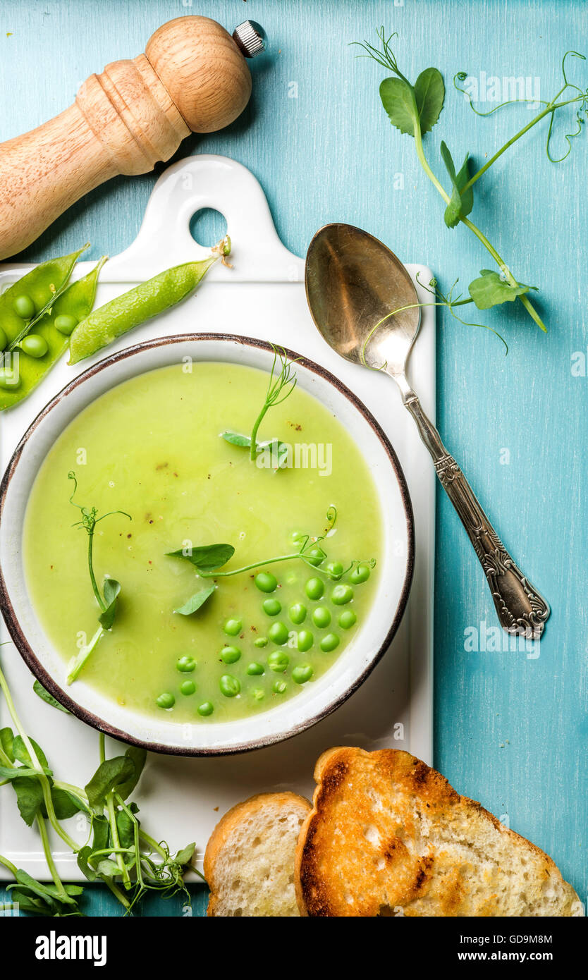 Luce estate pisello verde minestra in crema in tazza servita con i germogli, crostini di pane e spezie in ceramica bianca scheda sopra il turchese blu Foto Stock