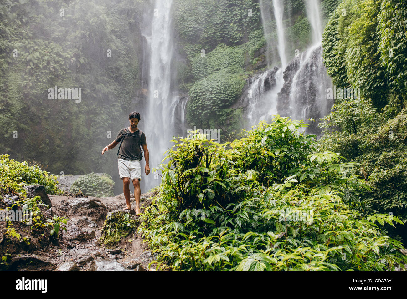 Colpo all'aperto del giovane uomo a camminare lungo il sentiero di montagna. Escursionista maschio escursioni nella foresta con cascata in background. Foto Stock