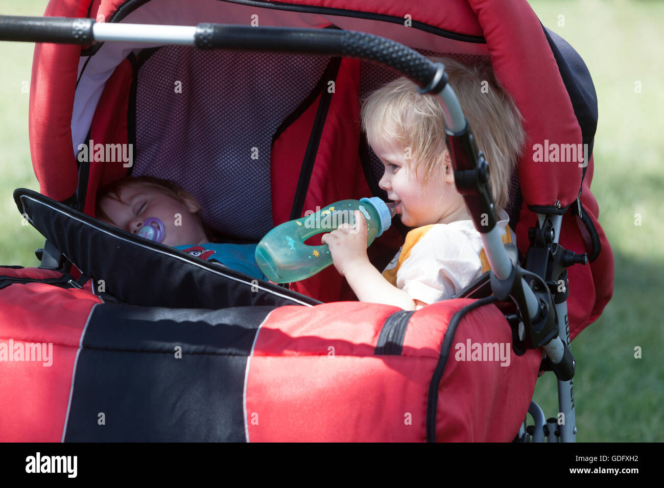 Bambini, gemelli in un passeggino, estate calda, un bambino beve da una tazza da biberon Foto Stock