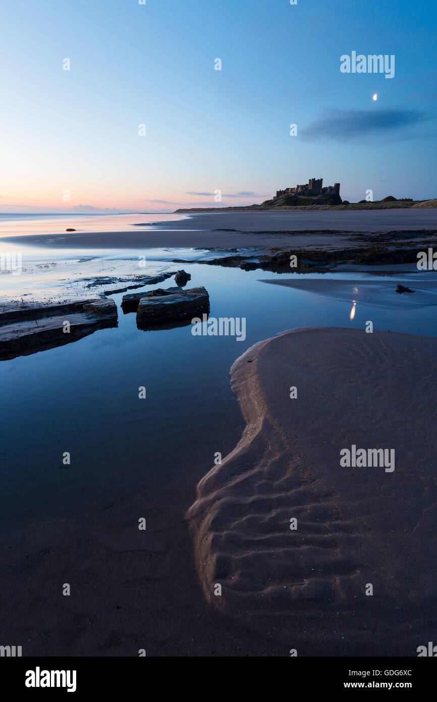 Luna oltre il castello di Bamburgh all'alba Foto Stock