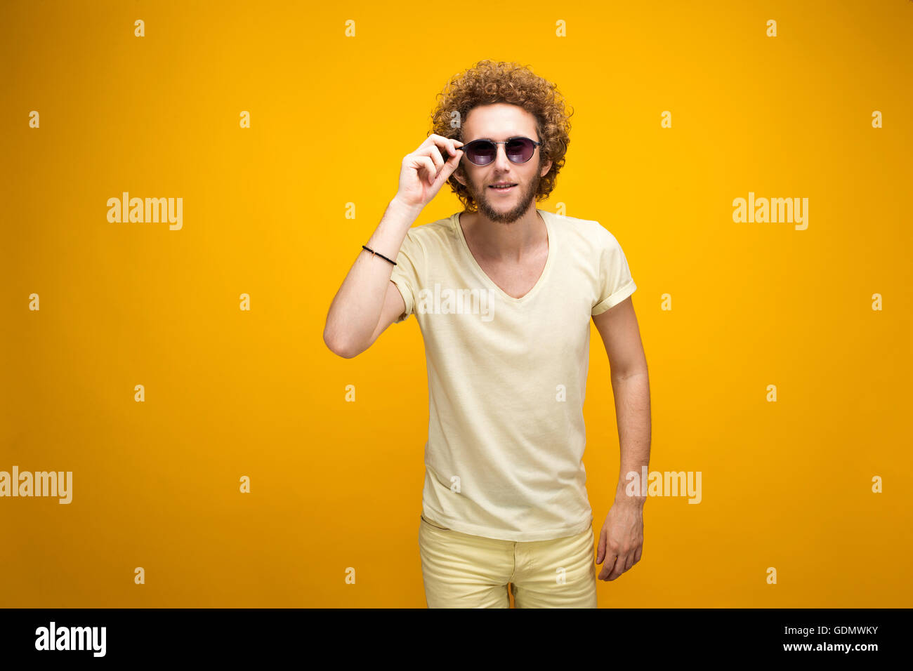 Ritratto di curly-pelose giovane uomo in occhiali da sole e bermuda cercando confuso a telecamera. Isolare. Sfondo giallo. Foto Stock