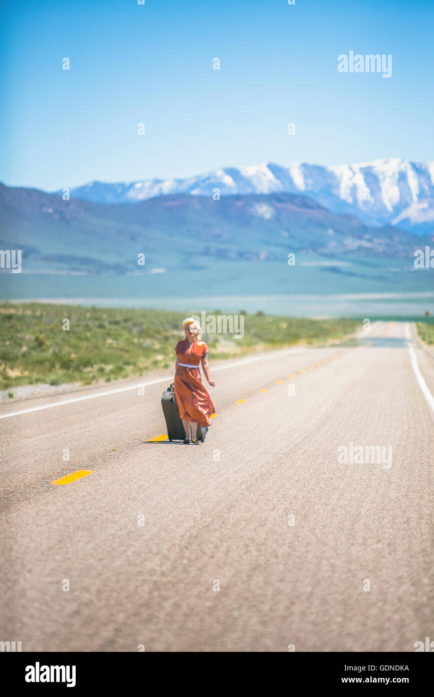 I giovani degli anni cinquanta donna stile camminare da solo sulla Highway 50 tirando la valigia su ruote, Nevada, STATI UNITI D'AMERICA Foto Stock