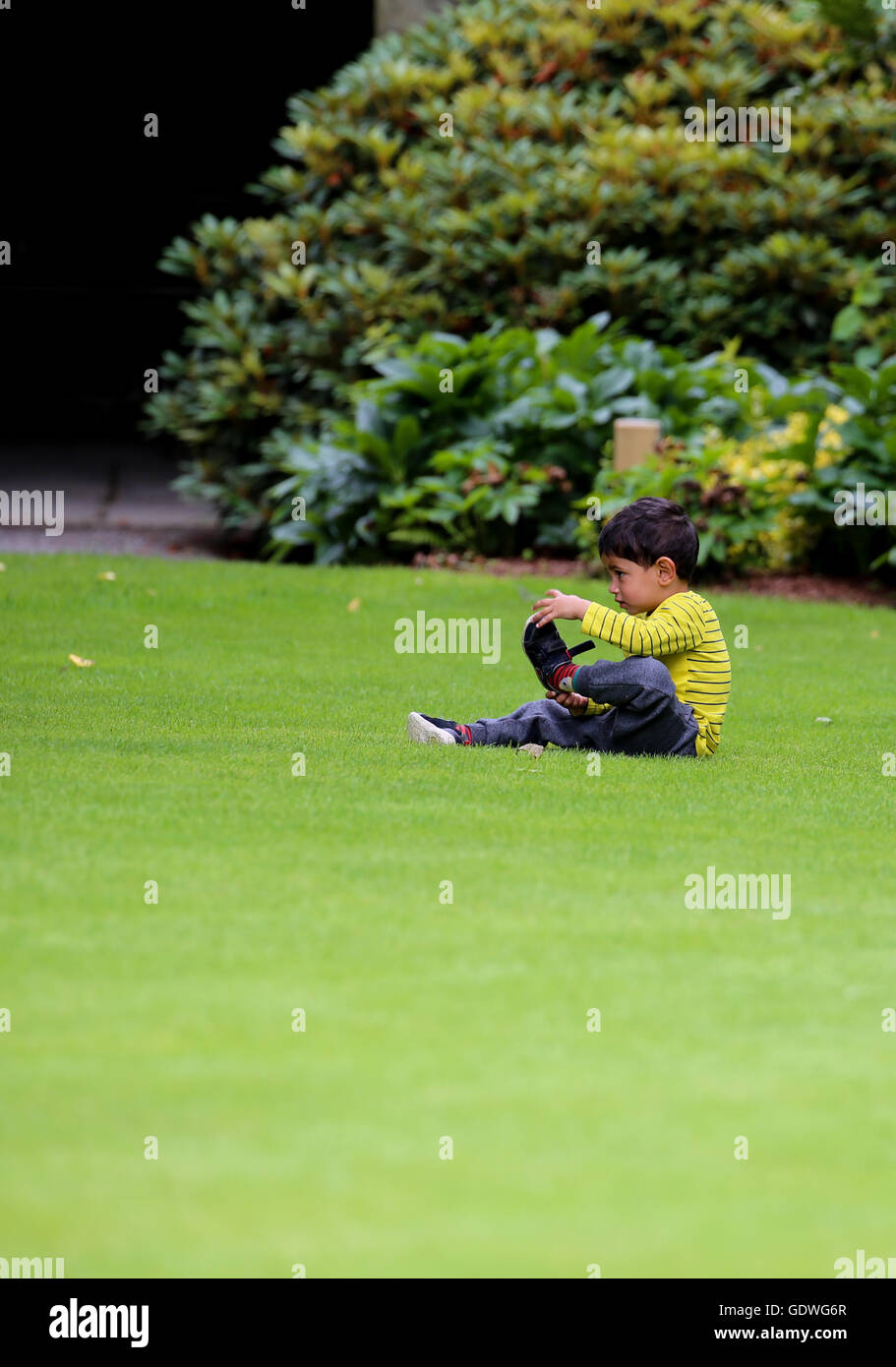 Piccolo Ragazzo tenendo le sue scarpe off sull'erba in un giardino, i jeans e un giallo listati top. Foto Stock