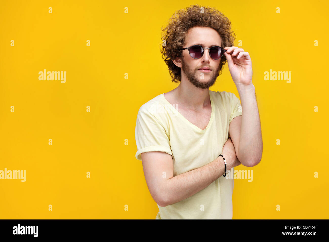 Ritratto di curly-pelose giovane uomo in occhiali da sole e bermuda cercando confuso a telecamera. Isolare. Sfondo giallo. Foto Stock