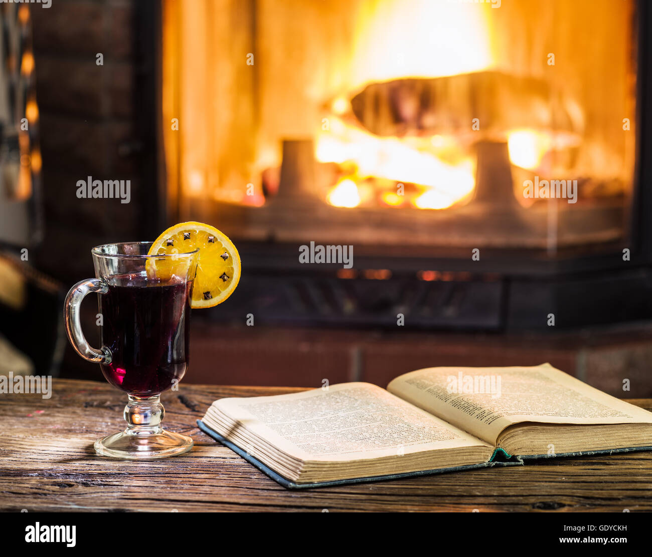 Vin brulé e un libro sul tavolo di legno. Camino con fuoco caldo sullo sfondo. Foto Stock