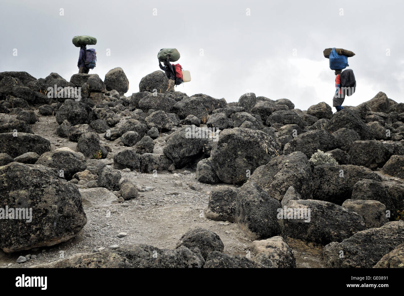 Tre i facchini che trasportano i sacchi di grandi dimensioni sulla loro testa e grandi rocce vulcaniche, Monte Kilimanjaro National Park, Tanzania Foto Stock
